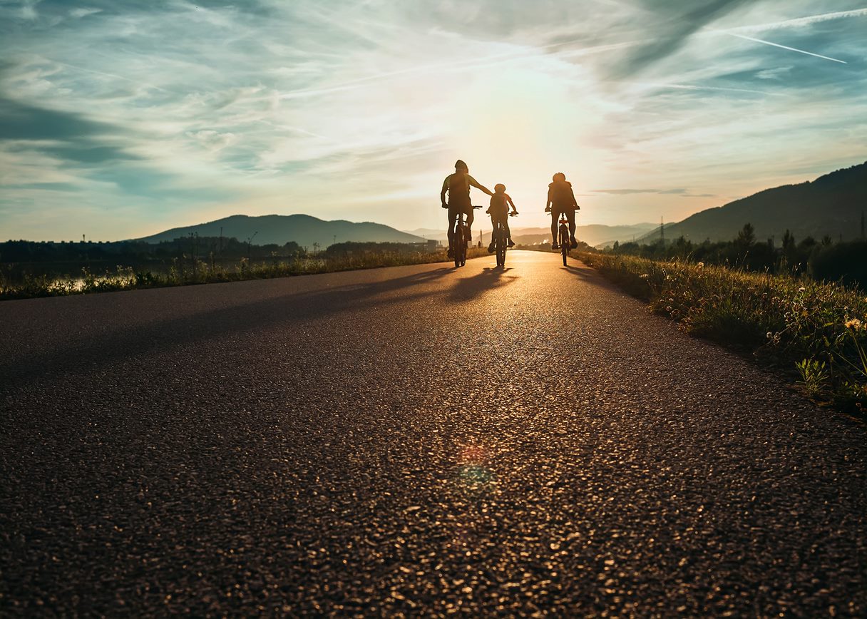 Three people cycle along the road, in the countryside at sunset.