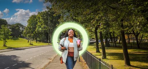 woman walking on a pavement next to a park with tress, holding a tablet and a pink travel mug