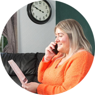 woman in orange shirt, sitting on a black sofa, speaking on a mobile and looking at a pamphlet