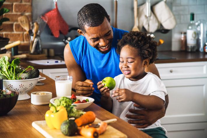 A man and child at the kitchen table with a chopping board of food in front of them, and the child is holding an apple.