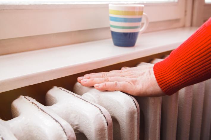 A person in a red jumper placing their hand on a white radiator, with a stripey sitting on the windowsill.