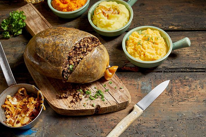 A table with a cut open haggis on a chopping board, surrounded by small bowls of potato and turnips