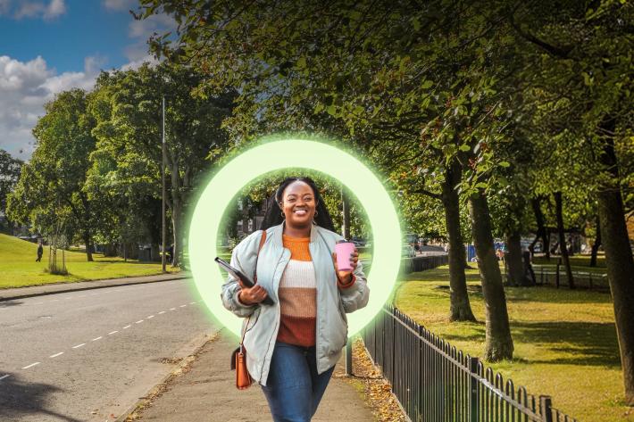 woman walking on a pavement next to a park with tress, holding a tablet and a pink travel mug
