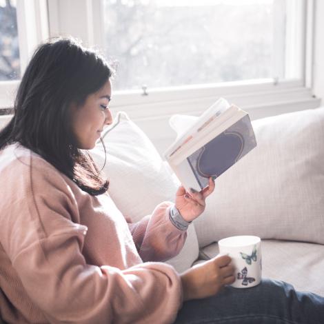 A woman sitting on a couch by a window, reading a book and holding a cup of tea.