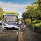 A family walks off a bus onto the pavement of a leafy street.