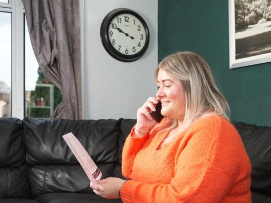 woman in orange shirt, sitting on a black sofa, speaking on a mobile and looking at a pamphlet